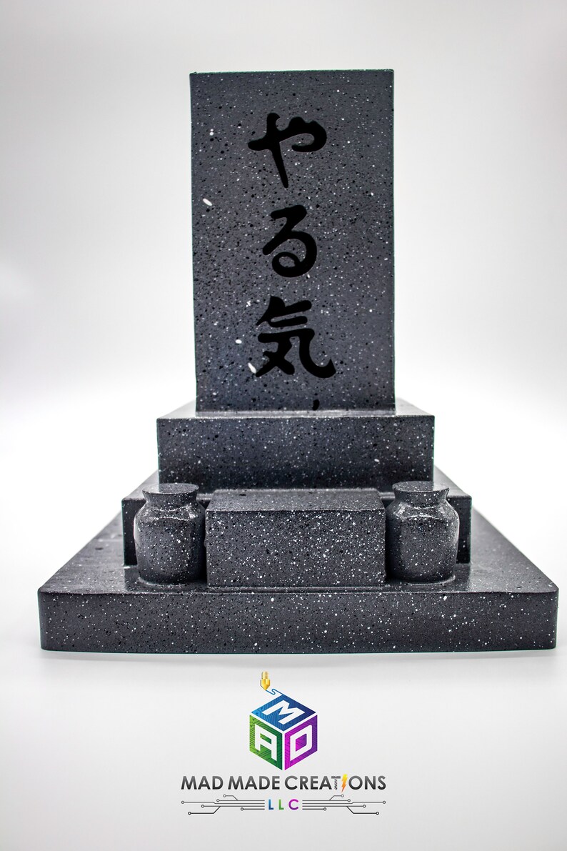 May include: A black granite Japanese style grave marker with the inscription "やる気" in black lettering. The marker is made up of three tiers with two small black urns on the bottom tier.