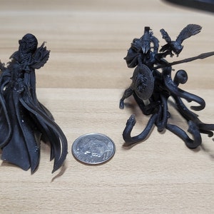 May include: Two black 3D printed figurines, one of a cloaked figure and the other of a creature with wings and a spear, are shown next to a US quarter for scale.