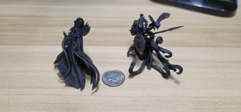 May include: Two black 3D printed figurines, one of a cloaked figure and the other of a creature with wings and a spear, are shown next to a US quarter for scale.