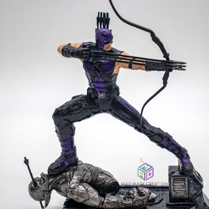 May include: A purple and black action figure of a superhero holding a bow and arrow. The figure is standing on a fallen figure with an arrow through its head. The base of the figure has the text "MAD MADE CREATION".