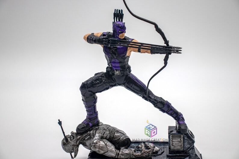 May include: A purple and black action figure of a superhero holding a bow and arrow. The figure is standing on a fallen figure with an arrow through its head. The base of the figure has the text "MAD MADE CREATION".