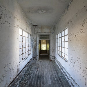 Puede incluir: Un pasillo interior con pintura blanca descascarillada y m&uacute;ltiples ventanas. Las tablas del suelo de madera est&aacute;n desgastadas, y la escena sugiere un edificio abandonado. El pasillo conduce a una puerta al final, con luz solar visible.