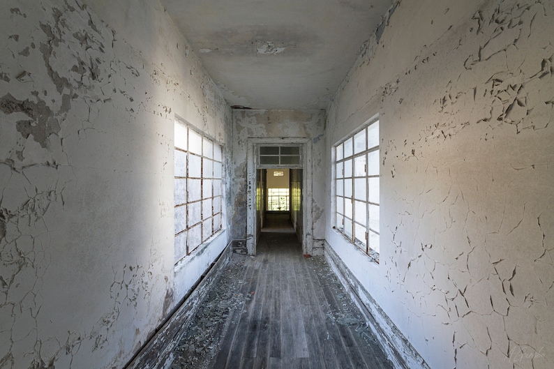 Puede incluir: Un pasillo interior con pintura blanca descascarillada y m&uacute;ltiples ventanas. Las tablas del suelo de madera est&aacute;n desgastadas, y la escena sugiere un edificio abandonado. El pasillo conduce a una puerta al final, con luz solar visible.