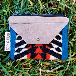 May include: A small, blue, red, white, and black patterned wallet with a gray suede flap and a zipper closure. The wallet has a tag that says "Scenic Route".