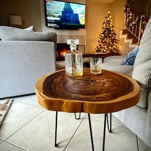 May include: A live edge wood coffee table with black hairpin legs. A crystal decanter and a rocks glass sit on the table. The table is in a living room with a lit Christmas tree and a fireplace.