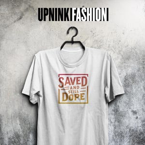 Peut inclure: T-shirt blanc avec l'inscription "SAVED AND STILL DOPE" dans un dégradé de rouge et d'or. Le t-shirt est suspendu à un cintre noir sur un fond gris texturé. La marque "UPNINK FASHION" est en haut.
