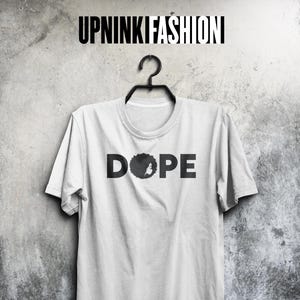Peut inclure: T-shirt blanc avec le mot "DOPE" en lettres noires. Le "O" est remplacé par une silhouette noire d'une tête de personne avec une afro. Le t-shirt est suspendu à un cintre noir contre un mur gris texturé.