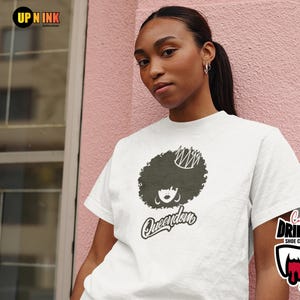 May include: White t-shirt with a black graphic of a woman's afro, crown, and earrings, with the word "Queendom". The shirt is worn by a person against a pink wall. The image also has logos.