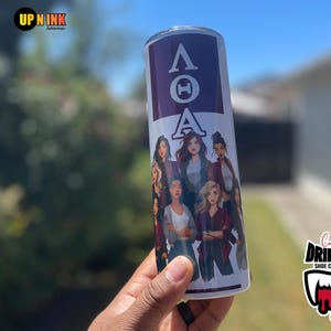 May include: A white tumbler with a purple top section featuring the Greek letters ΛΘA. The tumbler has a design of several women in various outfits. The tumbler is held by a person.