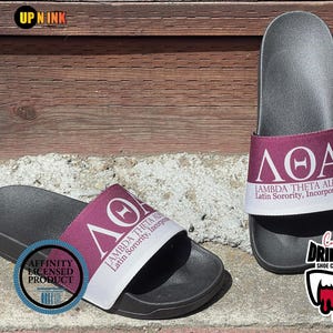 May include: Black slide sandals with a maroon and white strap featuring the Greek letters ΛΘΑ and the words "LAMBDA THETA ALPHA Latin Sorority, Incorporated." The sandals are an Affinity Licensed Product.