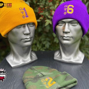 May include: Three beanies on display. A yellow beanie with purple text and a purple beanie with gold text are on mannequin heads. A camouflage beanie with gold text is at the bottom. The text on the beanies includes Greek letters and numbers.