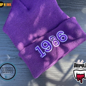 May include: A purple knitted beanie hat featuring the year "1986" embroidered in white and purple. The hat has a folded brim. The image also includes logos for "UP N INK", "Affinity Licensed Product", and "Count DRIPULA Shoe Customs".