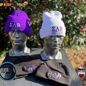 Puede incluir: Dos gorros de punto sobre cabezas de maniquí, uno morado con bordado blanco que dice ΣΛΒ ACE y uno blanco con bordado morado que dice ΣΛΒ DEUCE. Dos gorros negros debajo presentan bordados en morado y blanco que dicen ΣΛΒ TRE.
