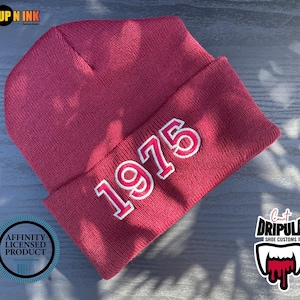 May include: A maroon knitted beanie hat featuring the year "1975" embroidered in white. The folded brim displays the year. The hat is made of a soft, knitted material.