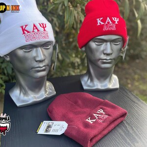 May include: Three beanies on display. A white beanie with red text reading "KAY Ψ DEUCE", a red beanie with white text reading "KAY Ψ ACE", and a maroon beanie with white text reading "KAY Ψ ACE".