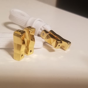 May include: Three gold-colored metal shoe lace aglets. The aglets are shaped like a pyramid with a small hole at the top. The aglets are on a white background.