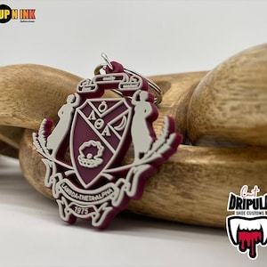 May include: A maroon and white Lambda Theta Alpha keychain with a shield design, featuring the Greek letters and a crown. The keychain has a silver keyring and the year 1975 is printed on the bottom.