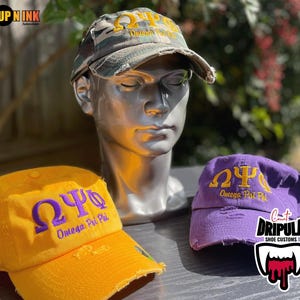 May include: Three baseball caps with distressed visors. One is yellow with purple embroidery, another is purple with gold embroidery, and the third is camouflage with gold embroidery. Each cap features the Greek letters Omega, Psi, and Phi, and the words "Omega Psi Phi".