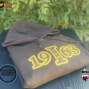 May include: A brown hooded sweatshirt with the number "1963" printed in yellow. The sweatshirt is folded on a dark surface, with green foliage in the background. The sweatshirt has a drawstring and a hood.
