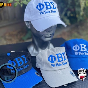 May include: Three baseball caps featuring the Phi Beta Sigma Greek letters and text. One cap is white with blue embroidery, one is gray with blue embroidery, and one is black with a blue brim and blue embroidery. Each cap has a distressed brim.