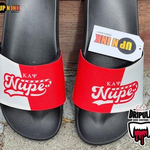 May include: A pair of black slides with a white and red design. The design features the text "K.A.Ψ Nupe" in a red and white font. The slides are a popular choice for sorority members and fans.