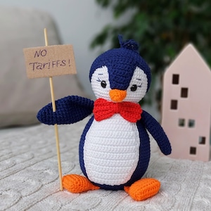 May include: A crocheted penguin toy holding a sign that says "NO TARIFFS!" The penguin is wearing a red bow tie and has orange feet. The penguin is sitting on a white knitted blanket.