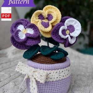 Può includere: Schema a uncinetto per una pianta in vaso con tre violette viola, bianche e gialle. Il vaso è viola con un nastro di pizzo bianco.