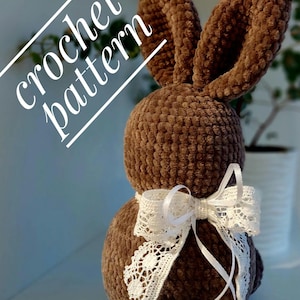 May include: A brown crocheted bunny with a white lace bow. The bunny is standing on a white surface. The text "crochet pattern" is written in white on a black background.
