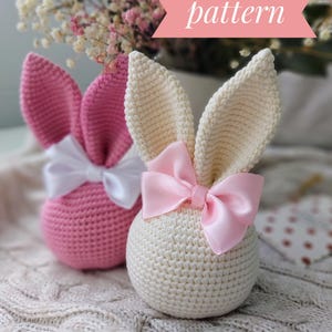 Könnte beinhalten: Zwei gehäkelte Hasen-Ornamente, eines rosa und eines cremefarben, mit Satinbändern. Die Ornamente liegen auf einer weißen Strickdecke. Häkelmuster.