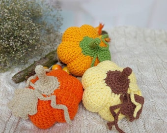 May include: Three crocheted pumpkins in orange, yellow, and brown with green stems. The pumpkins are sitting on a white knitted blanket.