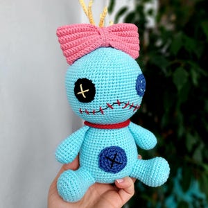 May include: A blue crocheted plush toy with a pink bow on its head. The toy has black eyes with a white cross, a red collar, and a blue button with a white cross on its chest. The toy has stitches on its face and body.