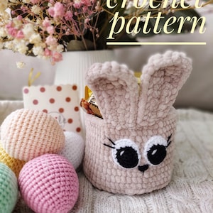 May include: Crochet pattern for a pink bunny-shaped basket with black eyes and white eyelashes. The basket is filled with small crocheted balls in various colors.