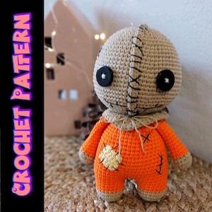 Op de afbeelding: Een handgemaakte gehaakte pop in een oranje jumpsuit met een bruin hoofd en gestikte details. De pop heeft grote zwarte knoopogen en een bruine, jute-achtige hoofdbedekking. De tekst "CROCHET PATTERN" wordt verticaal weergegeven.
