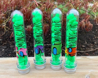 Puede incluir: Cuatro tubos de plástico transparente llenos de caramelos verdes. Cada tubo tiene una etiqueta que dice "Among Us" y presenta un personaje diferente del juego.