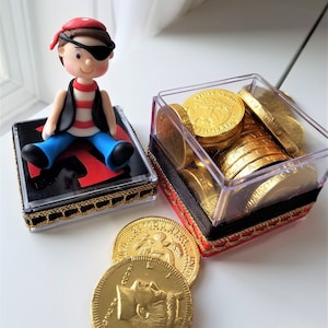 May include: A small pirate figurine with an eyepatch sits on a black and red box with gold trim. The figurine is holding a treasure chest filled with gold chocolate coins.