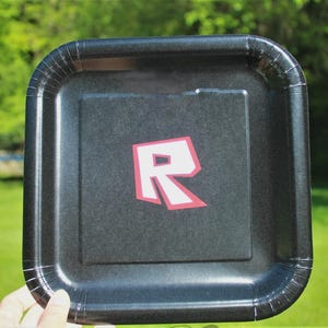 Pu&ograve; includere: Un piatto quadrato di carta nero con un logo "R" rosso e bianco. Il piatto ha una superficie leggermente strutturata e un bordo ondulato. Il logo &egrave; centrato.