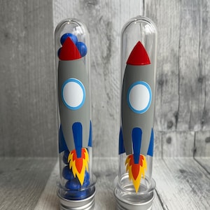 May include: Two clear plastic tubes with silver lids, each decorated with a gray and blue rocket ship design. The rockets have red noses and orange flames. The tubes are filled with blue and yellow candy.