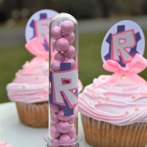 Pode incluir: Um tubo de pl&aacute;stico transparente cheio de doces cor-de-rosa, com uma tampa prateada e um adesivo "R" roxo e branco. O tubo est&aacute; sobre uma superf&iacute;cie branca ao lado de dois cupcakes cor-de-rosa com cobertura branca e granulado.