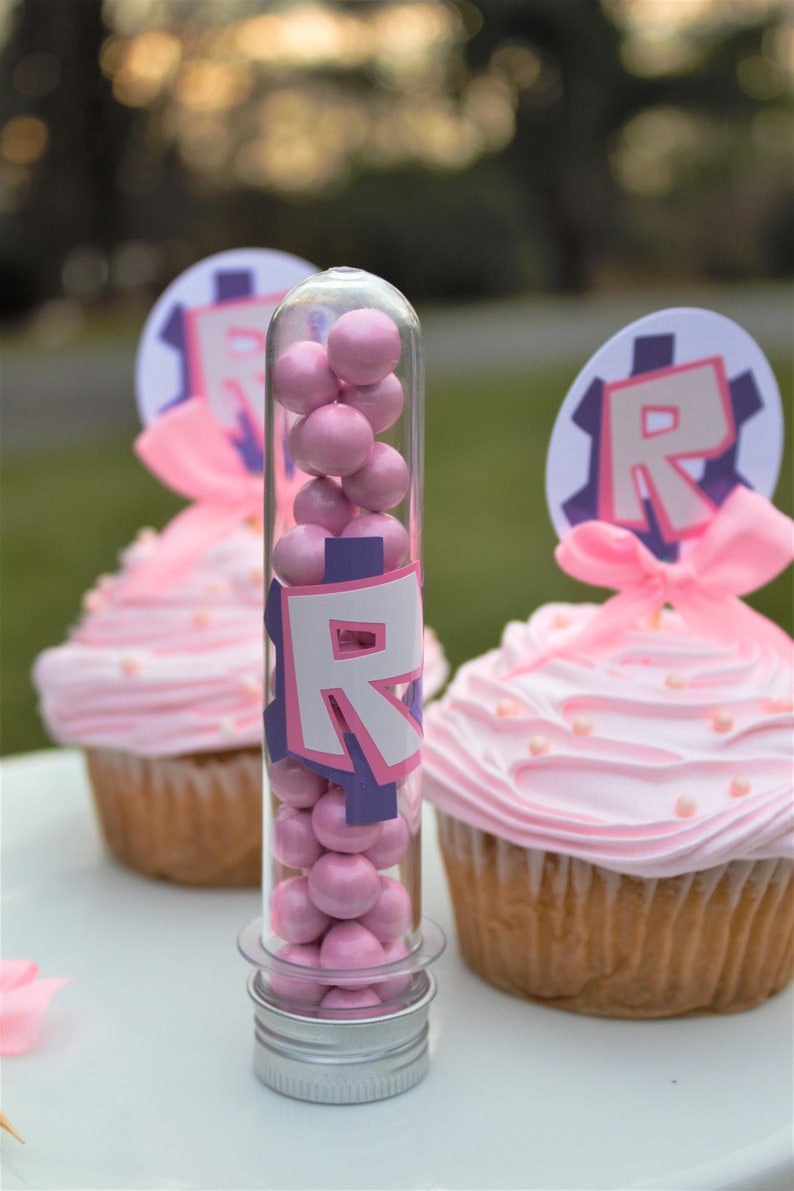 Pode incluir: Um tubo de pl&aacute;stico transparente cheio de doces cor-de-rosa, com uma tampa prateada e um adesivo "R" roxo e branco. O tubo est&aacute; sobre uma superf&iacute;cie branca ao lado de dois cupcakes cor-de-rosa com cobertura branca e granulado.