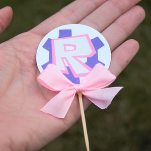 Pode incluir: Um la&ccedil;o rosa amarrado em torno de um palito de madeira com um c&iacute;rculo branco anexado. O c&iacute;rculo tem uma engrenagem roxa com uma letra "R" rosa dentro.