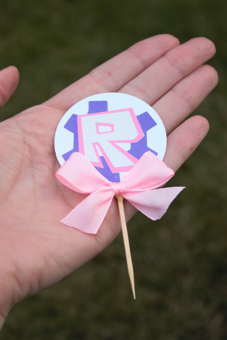 Pode incluir: Um la&ccedil;o rosa amarrado em torno de um palito de madeira com um c&iacute;rculo branco anexado. O c&iacute;rculo tem uma engrenagem roxa com uma letra "R" rosa dentro.