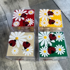 May include: Four clear plastic boxes with lids, each decorated with a different colored background and white daisies with red ladybugs. The boxes are filled with white candy.