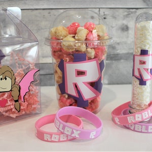 May include: A set of party favours featuring a clear plastic box filled with pink tissue paper and a pink and brown cartoon character with wings, a clear plastic cup filled with pink and white popcorn with a purple and white "R" on the side, a clear plastic tube filled with white candy with a purple and white "R" on the side, and two pink rubber bracelets with the word "ROBLOX" in white letters.