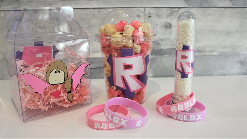 May include: A set of party favours featuring a clear plastic box filled with pink tissue paper and a pink and brown cartoon character with wings, a clear plastic cup filled with pink and white popcorn with a purple and white "R" on the side, a clear plastic tube filled with white candy with a purple and white "R" on the side, and two pink rubber bracelets with the word "ROBLOX" in white letters.