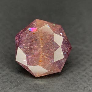 May include: A faceted, gemstone-like object with a complex geometric shape. The object has a reddish-purple hue with iridescent flashes of color. The surface reflects light, creating a sparkling effect. The gemstone is set against a dark background.