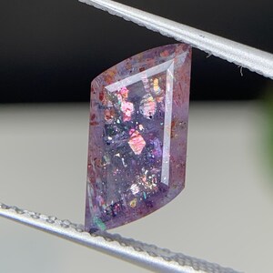 May include: A faceted gemstone held by tweezers. The gem is rectangular with angled edges. It has a deep purple colour with iridescent inclusions of pink, blue, and gold. The gem is held against a blurred background.