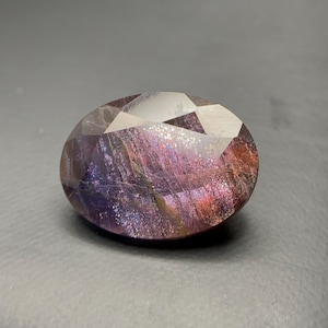 May include: An oval-shaped faceted gemstone displaying a deep purple and reddish-brown colour palette. The stone features internal inclusions that create a shimmering, iridescent effect with flashes of blue, gold, and violet light across its surface.
