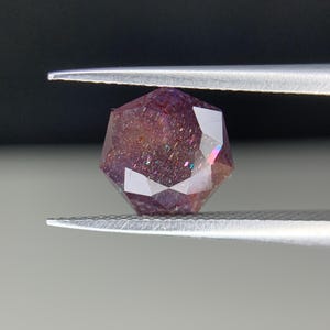 Op de afbeelding: Een gefacetteerde edelsteen, vastgehouden door een pincet, met een reeks kleuren. De edelsteen heeft een dieppaarse tint met iriserende flitsen van roze, blauw en groen. De snit is geometrisch, met meerdere facetten die het licht reflecteren.