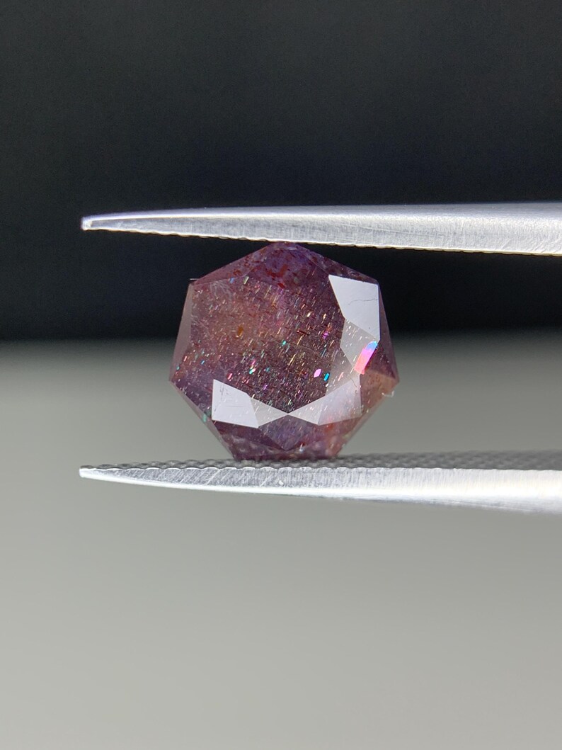 Op de afbeelding: Een gefacetteerde edelsteen, vastgehouden door een pincet, met een reeks kleuren. De edelsteen heeft een dieppaarse tint met iriserende flitsen van roze, blauw en groen. De snit is geometrisch, met meerdere facetten die het licht reflecteren.