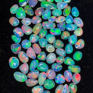 Puede incluir: Una colección de piedras preciosas de ópalo facetadas en varias formas y tamaños. Los ópalos muestran un vibrante juego de colores, con destellos de verde, azul, rojo y naranja. Las piedras están dispuestas sobre una superficie negra.
